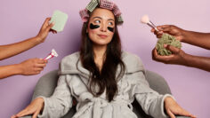 Relaxed young Korean woman in soft bathrobe, wears cosmetic patches under eyes, prepares for evening party, surrounded with many hands holding beauty items. Health care and wellbeing concept